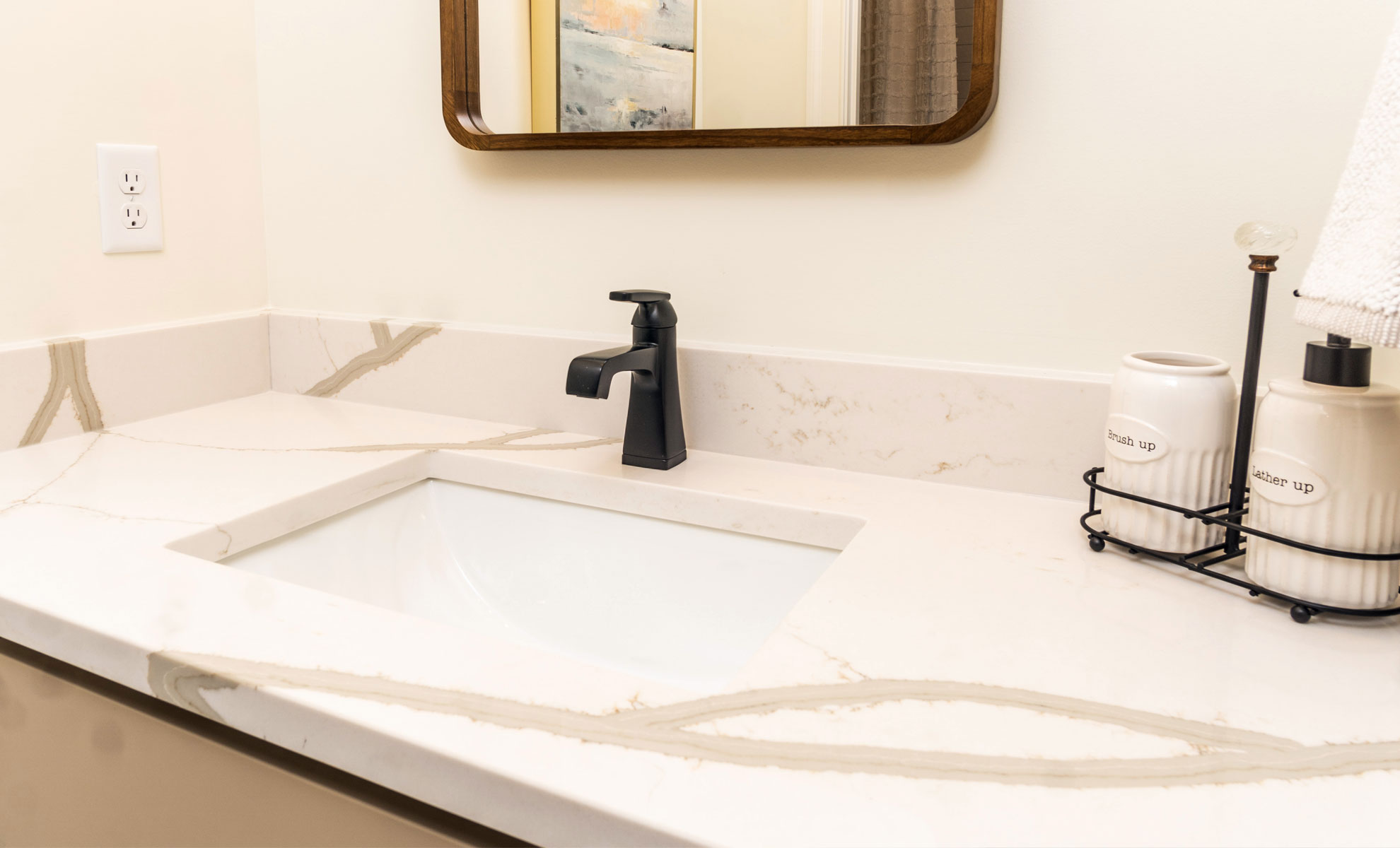 This image depicts a quartz bathroom countertop with thick veining and a white sink with black hardware. There is a soap dispenser next to the sink.