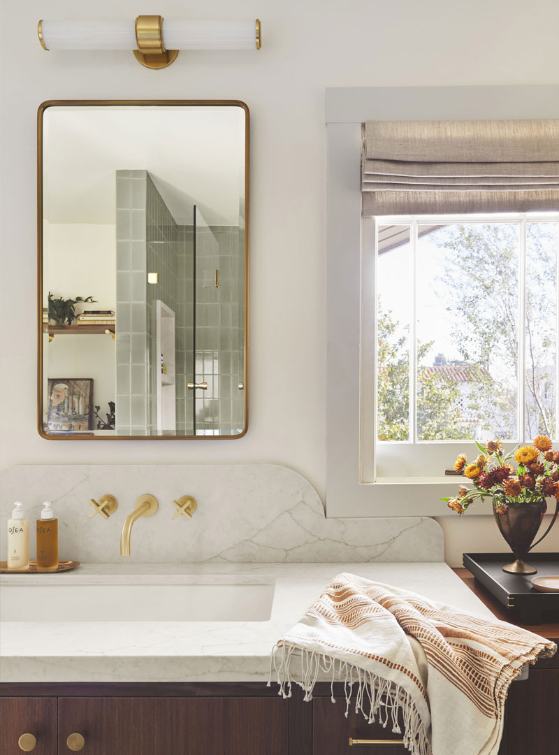 This image depicts a modern bathroom. Its sink and mirror feature gold hardware and a stunning white quartz countertop. There is a window to the right of the picture with open blinds and a vase of flowers next to it.