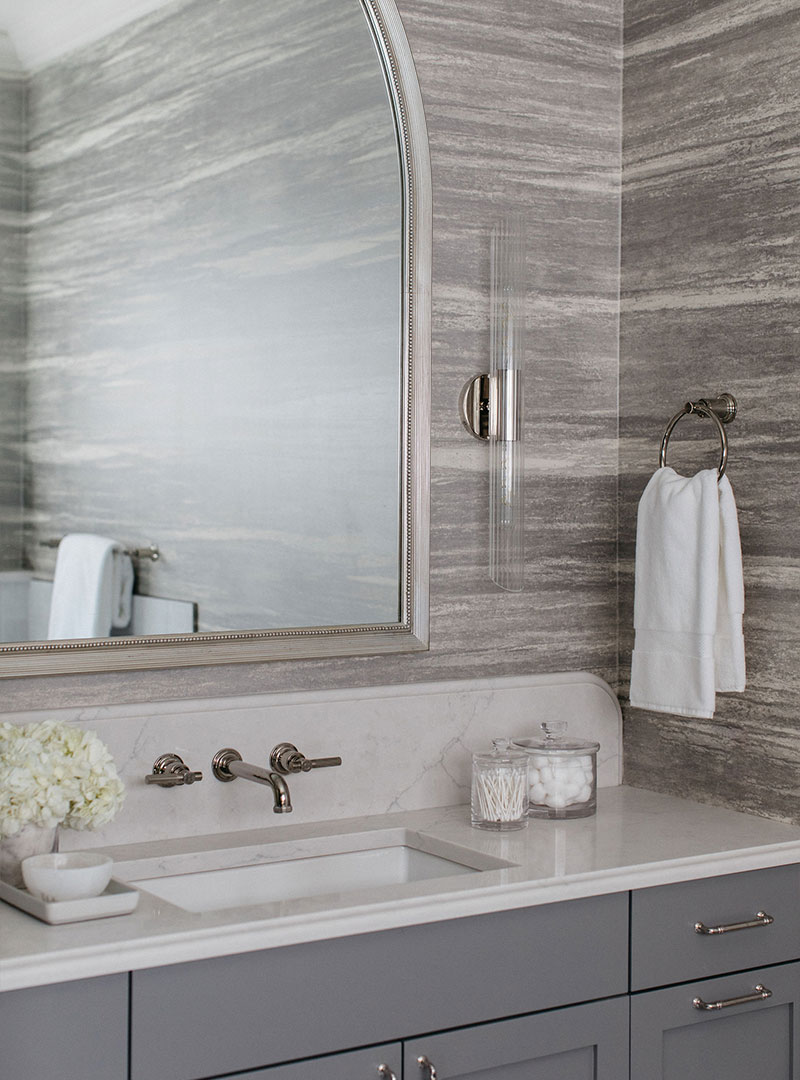 This image depicts a gray bathroom with a large mirror, a white handtowel hanging on the wall and a white quartz countertop. The sink’s hardware is silver, and there is a small vase of white flowers next to the sink. 