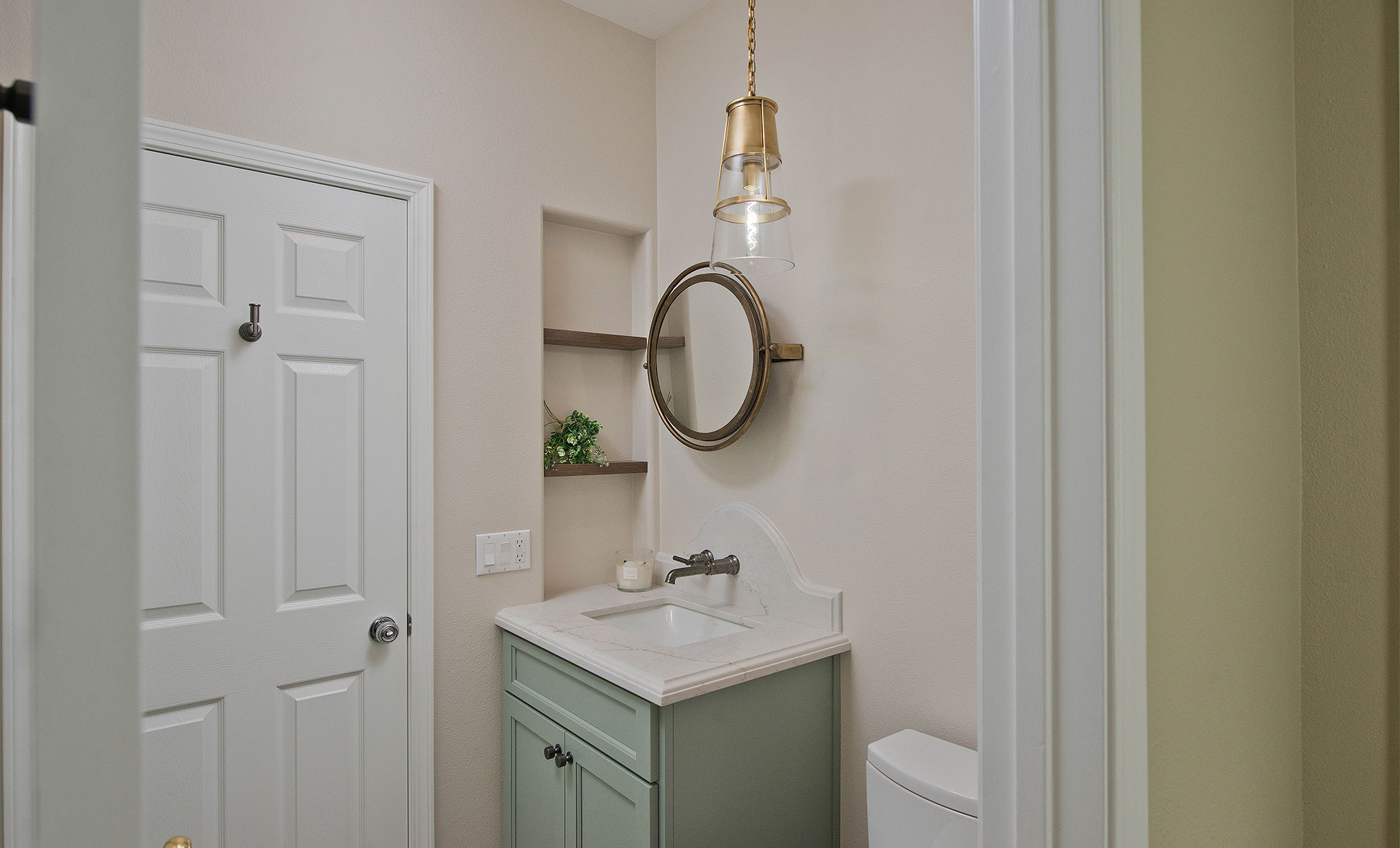 This image depicts a small bathroom with beige-colored walls and a white door. A small standalone sink with a white quartz countertop attached to a green cabinet sits next to a white toilet.