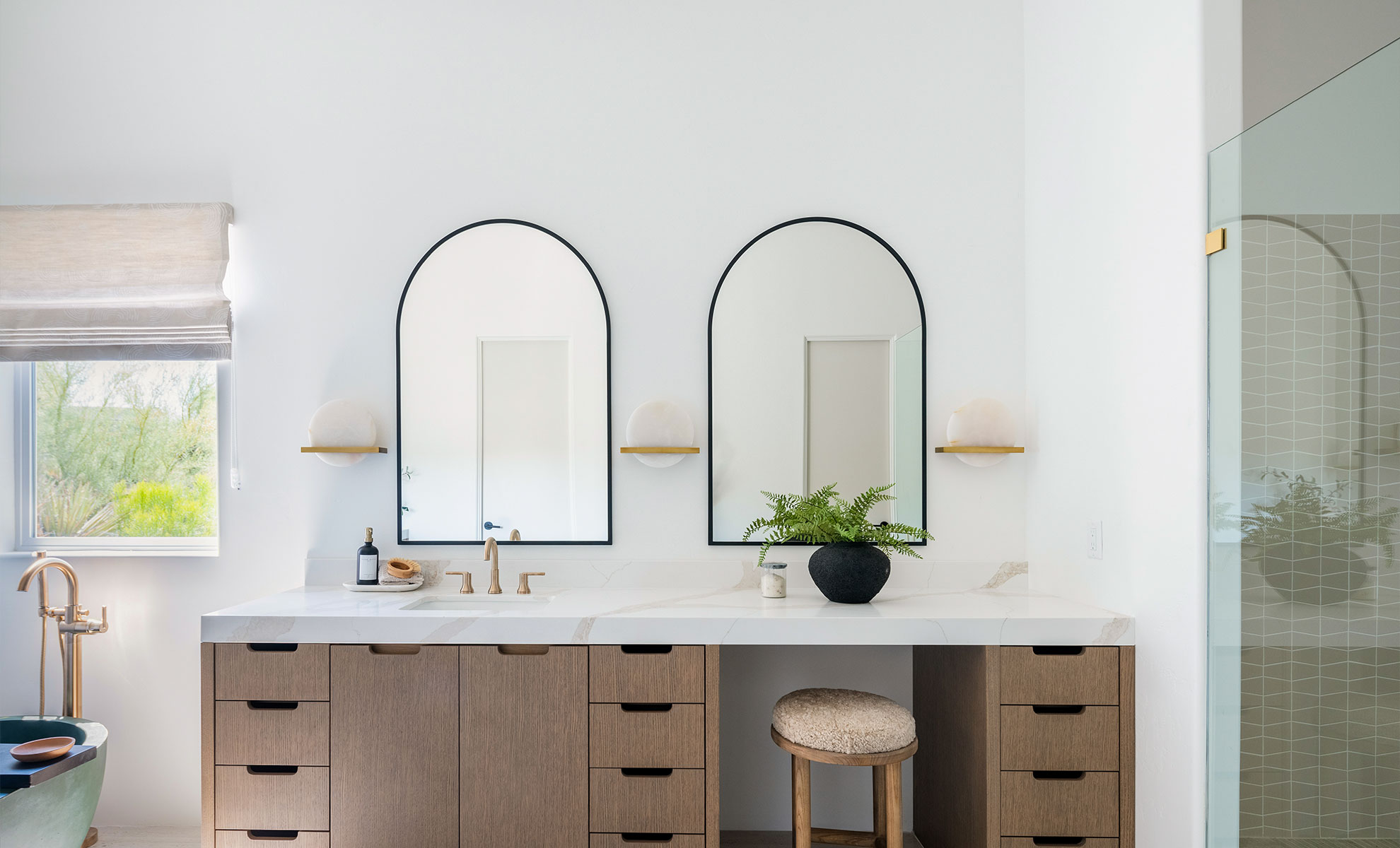 This image depicts a spacious bathroom with wooden cabinets, double sinks and a minimalistic quartz countertop. The sink hardware is gold. There are mirrors with black borders above the sink. 